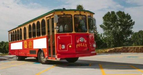 A red and yellow trolley bus named "Polly" parked in a scenic area with trees and blue skies in the background.