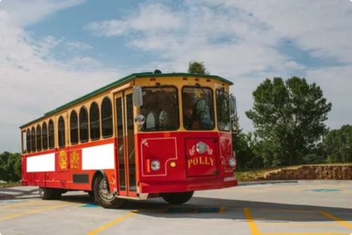 A red and yellow trolley bus named "Polly" parked in a lot with trees in the background under a cloudy sky.