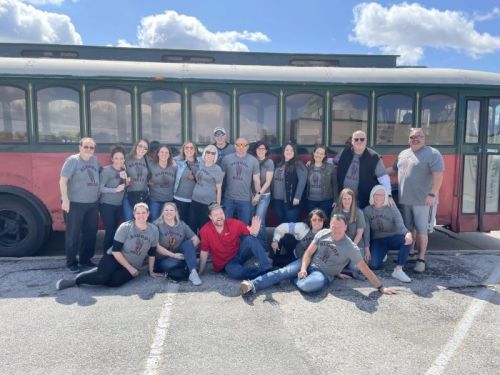 A group of people in matching gray shirts poses in front of a vintage bus on a sunny day.