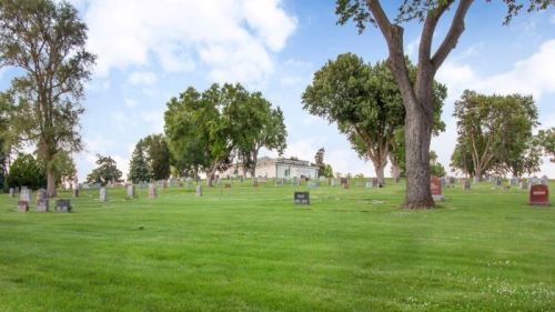 A peaceful cemetery with gravestones scattered across a green lawn, surrounded by trees and a blue sky.
