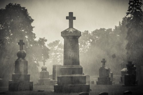 A misty graveyard with tall gravestones and crosses, shrouded in rain and fog, creating a somber atmosphere.