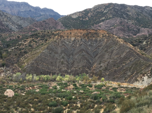 Geological formation with zigzag patterns on a hillside, surrounded by greenery and mountains in the background.