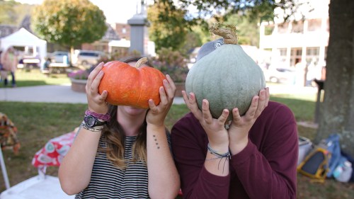 Two people hold pumpkins in front of their faces, smiling, at an outdoor market with trees and tents in the background.