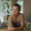 A person with tattoos sits thoughtfully at a table in a bright, modern living space.