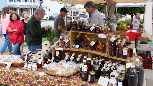 A bustling farmers market with vendors selling jars of honey and syrup, surrounded by shoppers and colorful displays.