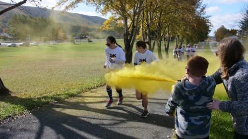 Runners in a 5K race pass through a cloud of yellow powder, with spectators cheering on the sidelines.