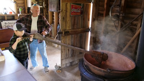 A child and an older man stir a large pot in a rustic workshop, with steam rising and vintage decor in the background.