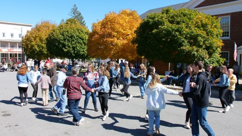 A lively outdoor dance event with people in casual attire dancing under vibrant autumn trees.