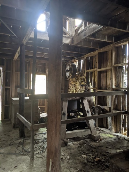 Interior of an old, rustic building with wooden beams, machinery, and scattered debris. Light filters through broken windows.