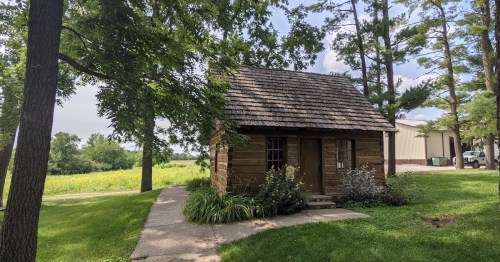 A small log cabin surrounded by trees and grass, with a path leading to the entrance and fields in the background.