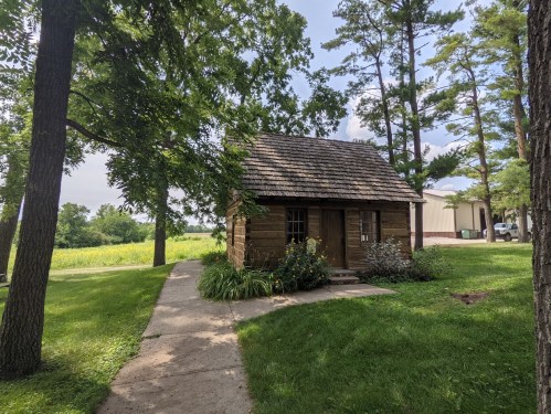 A small log cabin surrounded by trees and greenery, with a path leading to the entrance.