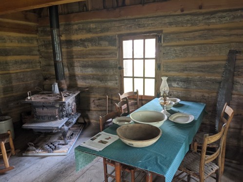 A rustic wooden room with a table set for a meal, a stove, and vintage kitchenware. Natural light through a window.