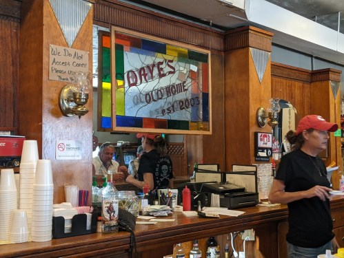 A bar interior with wooden decor, a stained glass sign reading "Dave's Old Home," and a woman in a red cap.