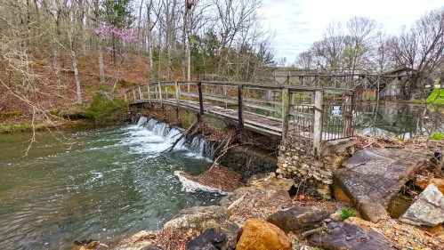 A wooden bridge over a stream with small waterfalls, surrounded by trees and rocky terrain in a serene landscape.