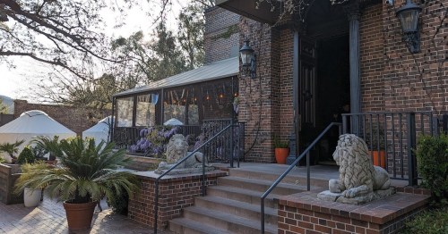 A brick building entrance with lion statues, steps, and outdoor seating surrounded by greenery and string lights.