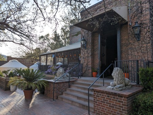 A brick building entrance with lion statues, steps, and outdoor seating surrounded by greenery and trees.