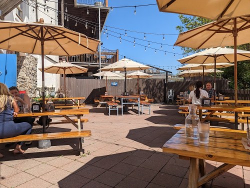 Outdoor dining area with wooden tables and umbrellas, string lights overhead, and a few patrons enjoying the space.
