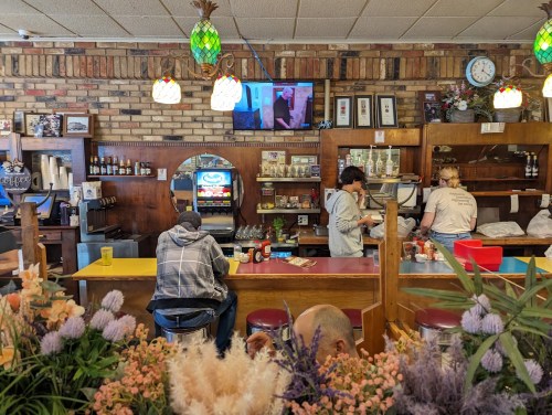 A cozy café interior with patrons at the counter, a TV on the wall, and colorful flowers in the foreground.