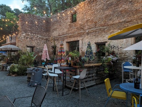 A rustic outdoor space with string lights, colorful umbrellas, and plants, featuring tables and chairs among stone walls.