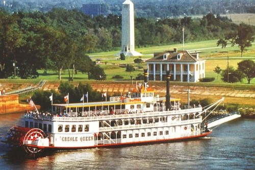 A steamboat named Creole Queen sails on a river, with a historic building and monument in the background.