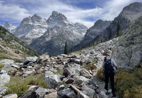 A hiker navigates a rocky trail with towering mountains and a blue sky in the background.