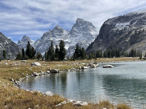 A serene mountain landscape with a clear lake, rocky shore, and towering snow-capped peaks under a cloudy sky.