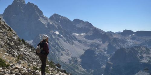 A hiker stands on a rocky mountain trail, gazing at a stunning mountain landscape under a clear blue sky.