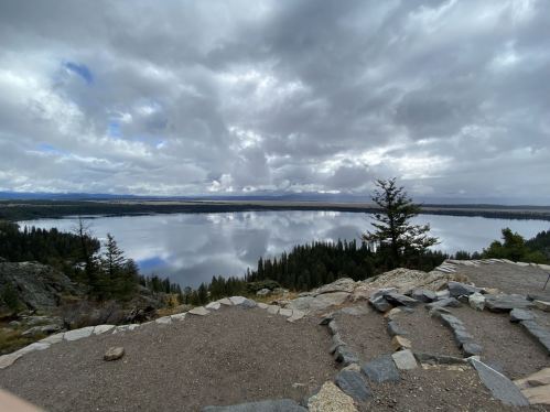 A serene lake reflects cloudy skies, surrounded by trees and rocky terrain.