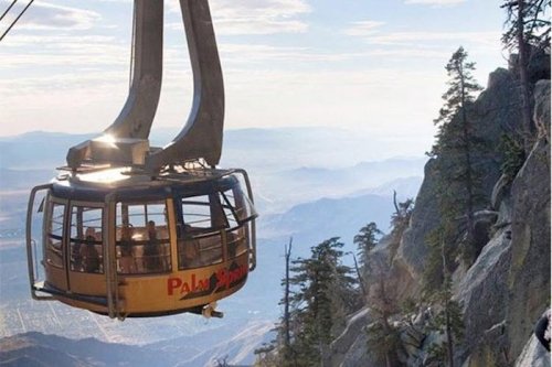 A gondola lifts passengers above a mountainous landscape, surrounded by trees and distant hills under a clear sky.