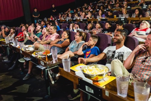A lively audience enjoying a movie in a theater, laughing and eating snacks, with drinks on their tables.