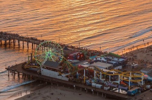 Aerial view of a vibrant pier with a ferris wheel at sunset, reflecting golden hues on the water.