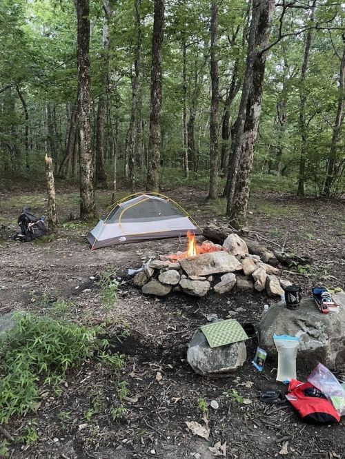 A campsite with a tent, a campfire surrounded by rocks, and scattered gear in a wooded area.
