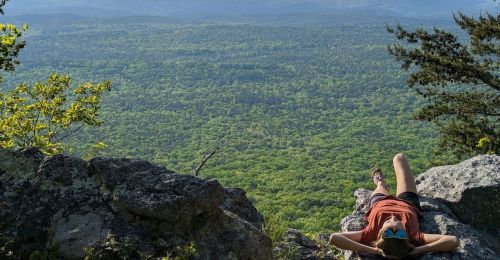 A person relaxes on a rocky ledge, overlooking a vast green landscape and distant mountains under a clear sky.