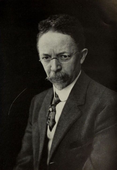 A serious man with glasses and a mustache, wearing a suit and tie, poses against a dark background.