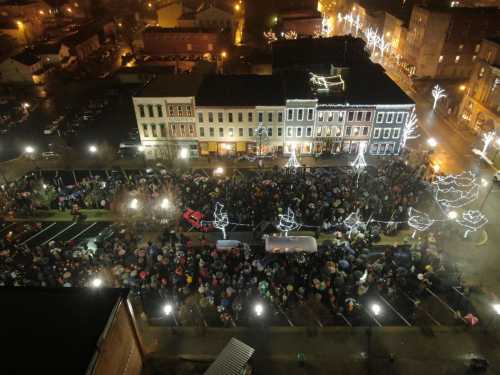 A large crowd gathers in a festive, illuminated town square, with holiday lights and umbrellas visible in the night.