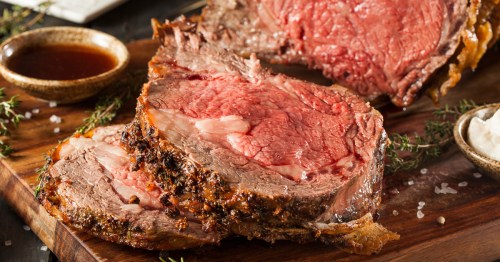 Tender slices of prime rib roast, seasoned with fresh herbs, served with a side of rich au jus on a rustic wooden board.