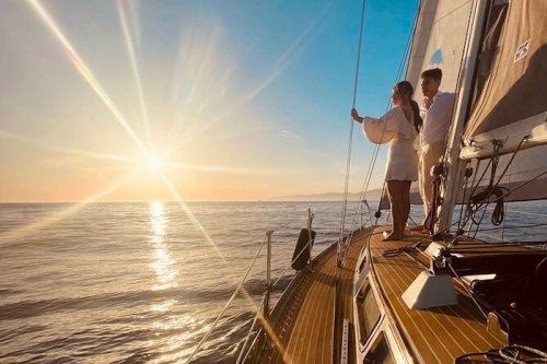 A couple stands on a sailboat at sunset, gazing at the horizon over calm waters. Sunlight sparkles on the waves.