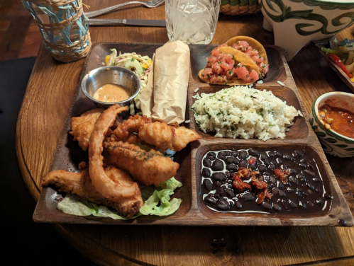 A wooden platter with fried fish, rice, black beans, a taco, salad, and dipping sauce.