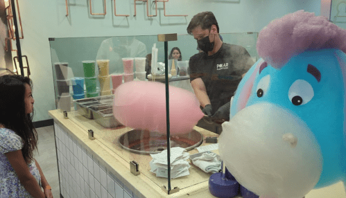 A child watches in awe as a worker makes cotton candy, with a large cartoon unicorn nearby.