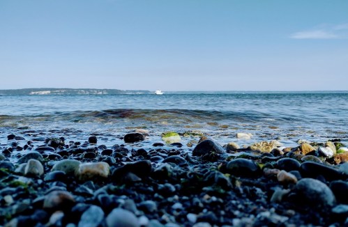 A rocky shoreline with smooth stones, gentle waves, and a distant boat on a calm sea under a clear blue sky.