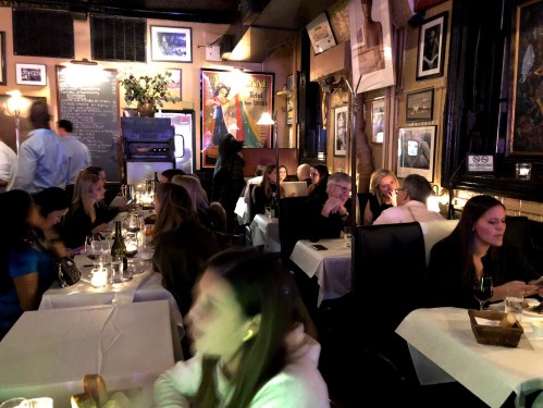 A bustling restaurant scene with diners at tables, dim lighting, and framed artwork on the walls.