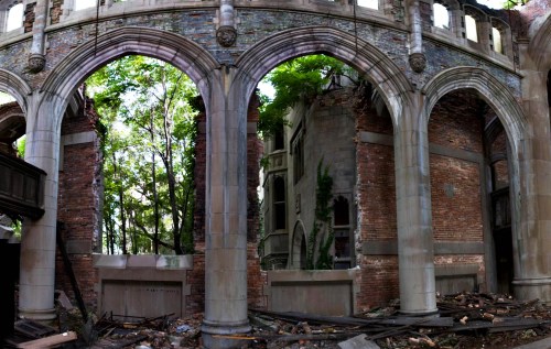 A panoramic view of a dilapidated building with arched stone walls, overgrown with greenery and debris on the floor.