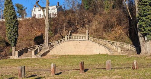 A stone bridge with decorative railings leads to a hillside, surrounded by trees and grass. A house is visible in the background.