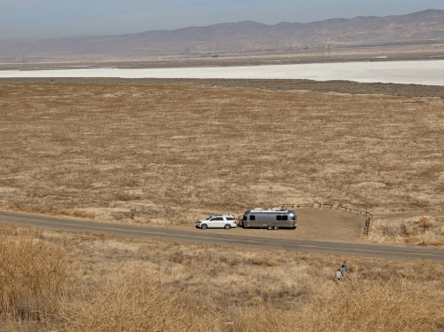A silver Airstream trailer parked beside a white vehicle on a dry, grassy landscape with distant mountains and salt flats.