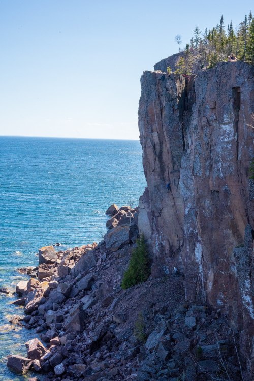 A rocky cliff overlooking a calm blue sea, with trees scattered along the top and a clear sky above.