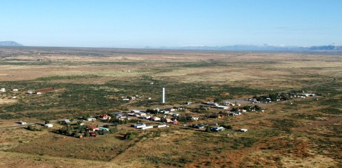 Aerial view of a small, rural town surrounded by vast, open landscapes and distant mountains under a clear blue sky.