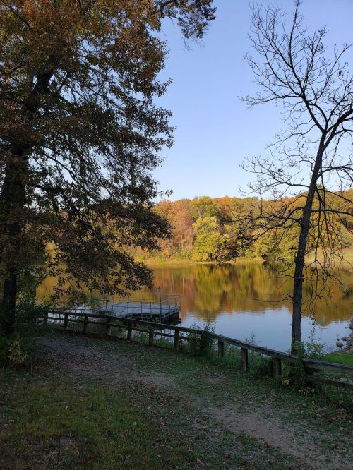A serene lake surrounded by trees, reflecting autumn colors under a clear blue sky. A wooden dock is visible.