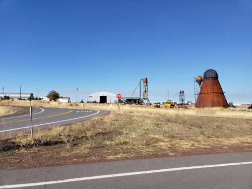 A rural scene featuring a winding road, a large metal structure, and industrial equipment against a clear blue sky.