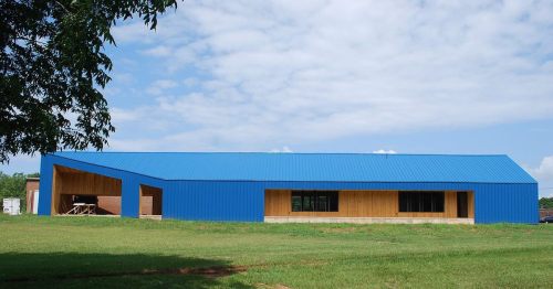 A modern building with a blue metal roof and wooden accents, set against a grassy landscape and cloudy sky.