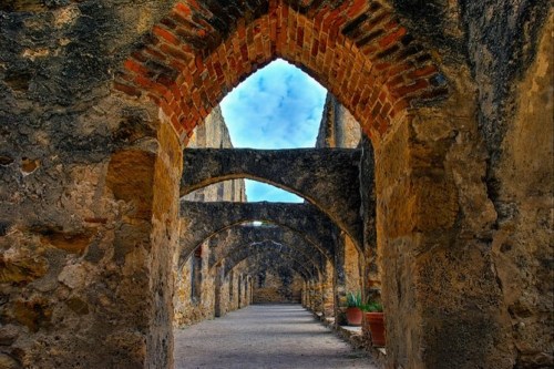 A stone corridor with arched brick ceilings, leading through ancient ruins under a blue sky.